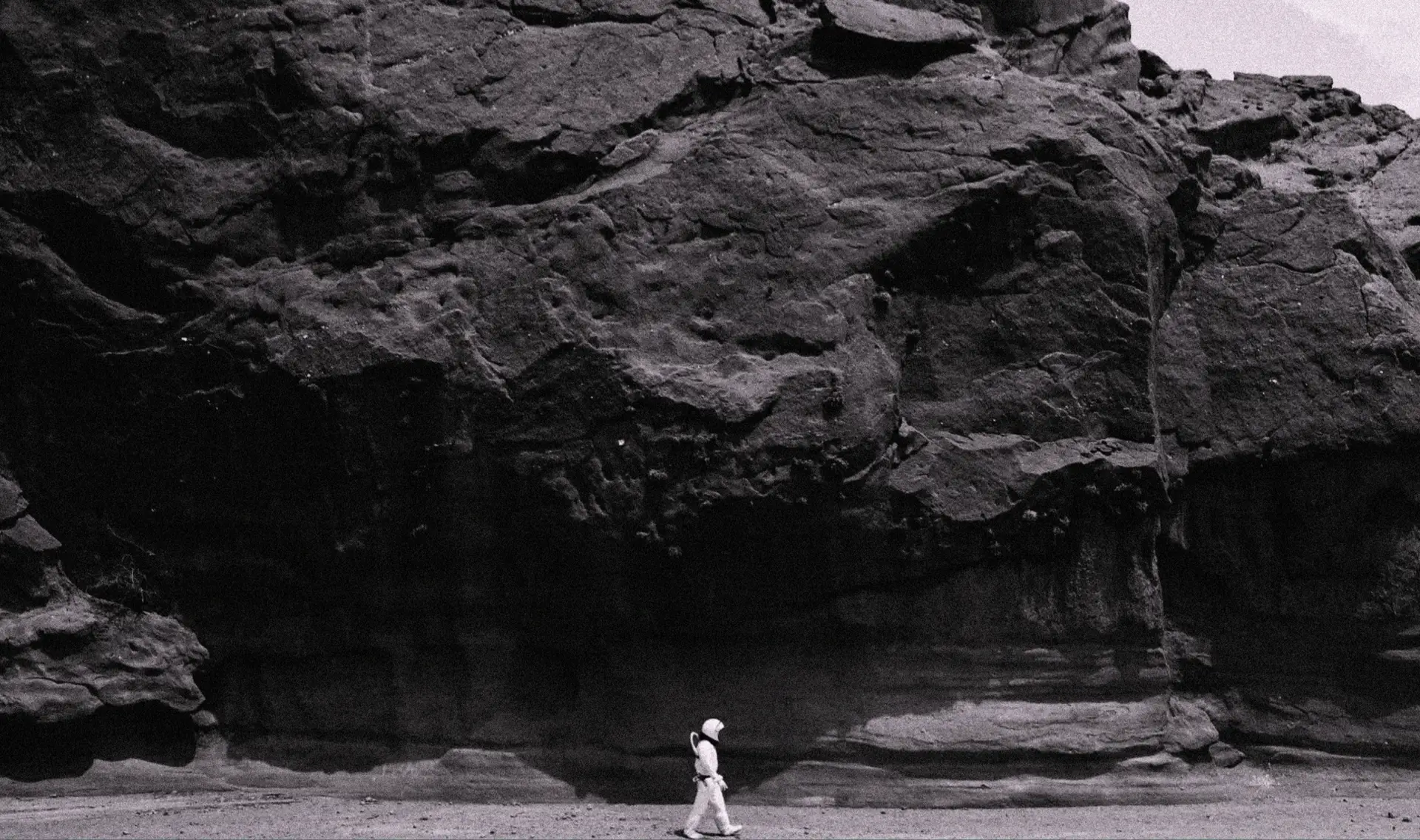 Black and white photo of astronaut walking along a massive rocky cliff, evoking the exploration and isolation of early Mars missions.