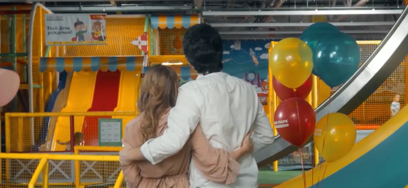 Parents with arms around each other watching their child play on slides at Chudo-Park family entertainment center, with balloons and birthday decorations.